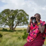 A Maasai guide in traditional red shuka using binoculars to track wildlife on the open savannah near Enkewa Camp.