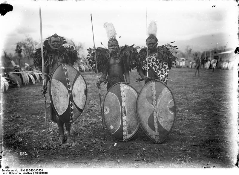 Black and white archival photograph from circa 1906 showing three Maasai warriors in full ceremonial dress — feathered headdresses, body paint, and decorated ox-hide shields — photographed in German East Africa.