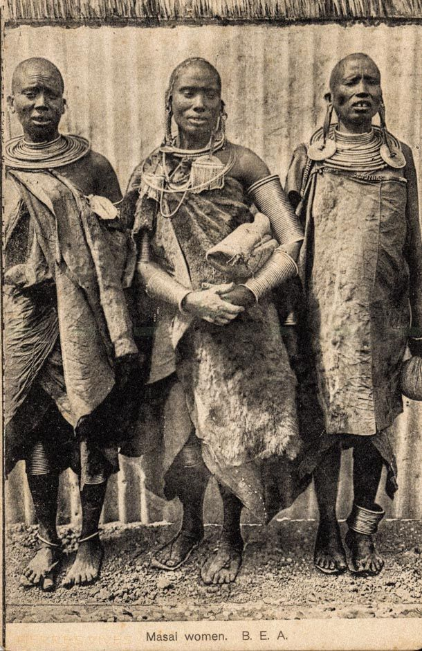 Sepia-toned archival postcard from British East Africa, circa 1900–1910, showing three Maasai women wearing traditional hide garments, layered coil necklaces, large disc earrings, and metal arm and leg bands.