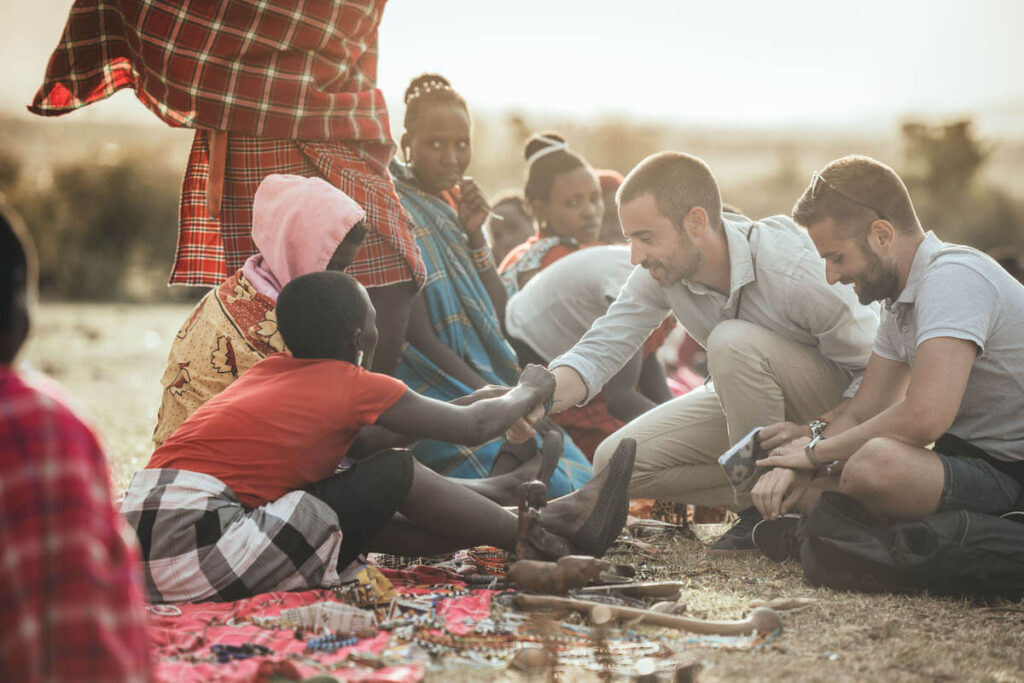 Two Enkewa Camp guests crouching down to greet a Maasai child during a guided village visit in the Olderkesi Conservancy, surrounded by Maasai women and traditional beadwork laid out on a cloth.